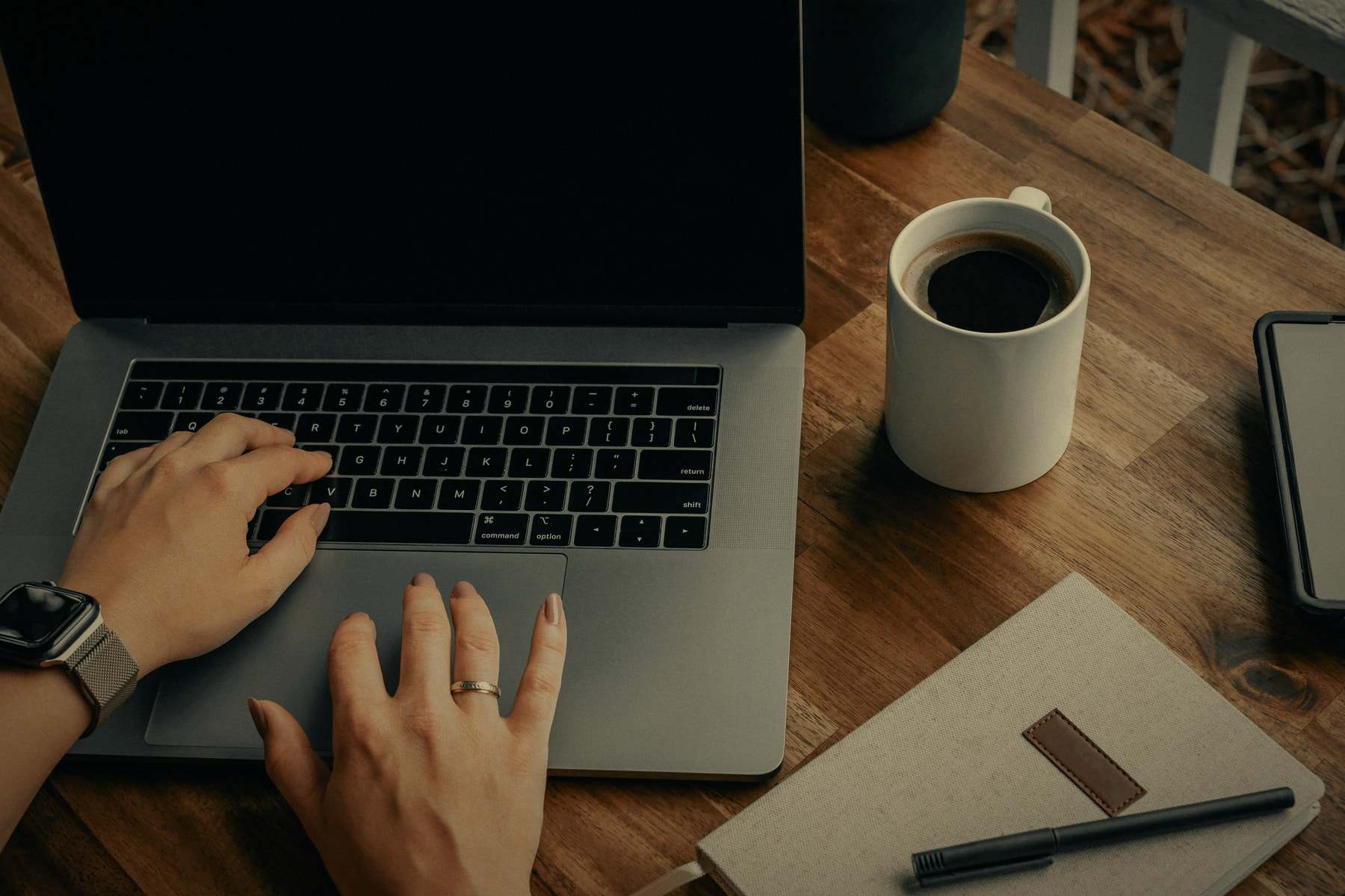 Mani al lavoro su un portatile, su un tavolo di legno con una tazza di caffè e un quaderno